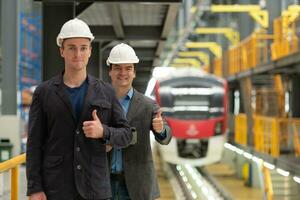 Porträt von zwei jung Ingenieure zeigen Daumen oben im Vorderseite von Zug Bahnhof foto