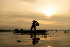 Silhouette von Fischer beim Sonnenaufgang, Stehen an Bord ein Rudern Boot und Gießen ein Netz zu Fang Fisch zum Essen foto
