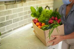 Nahansicht von Frau im medizinisch Maske, Tragen Karton Box mit frisch reif organisch gesund Gemüse, Früchte und Grüns foto