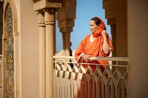 schön Frau im stilvoll orientalisch Kleidung und bedeckt Kopf im Schal von Orange Farbe, suchen in Distanz, im ein Moschee foto