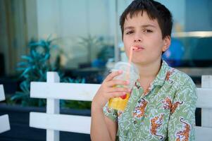 Porträt von ein gut aussehend kaukasisch Teenager Junge im Sommer- Kleider Trinken frisch gedrückt Saft von ein Stroh foto