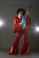 ein in voller Länge Mädchen im ein aufgeflammt Disco-Stil Bühne Kostüm hält ein Comic Gitarre im ihr Hände. foto