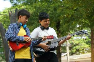 zwei asiatisch Jungs wurden haben Spaß spielen klassisch Gitarre zusammen während ihr kostenlos Zeit beim ein Schule Sommer- Lager beim das Park. Sanft und selektiv Fokus. foto