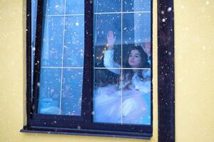 Mädchen sieht aus aus Fenster beim Schneefall von gemütlich festlich Zuhause draußen mit das warm Licht von Fee Beleuchtung Girlanden Innerhalb - - feiern Weihnachten und Neu Jahr im ein warm heim. foto