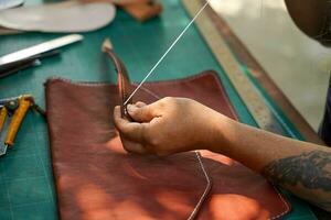 Nahansicht und Ernte Hände von Leder Handwerker Nähen ein Leder braun Tasche zum ein Kunde. foto