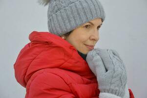 ein jung Frau im ein grau gestrickt Hut und warm Fäustlinge hält ihr Hände in der Nähe von ihr Gesicht. Porträt von ein kaukasisch Frau im ein rot Jacke mit ein Kapuze auf ein Licht Hintergrund. foto