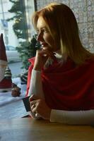 ein Mädchen mit blond Haar im ein rot Schal sitzt beim ein Fenster dekoriert zum Weihnachten. Porträt von ein jung Frau auf Weihnachten Vorabend, warten zum das Urlaub. foto