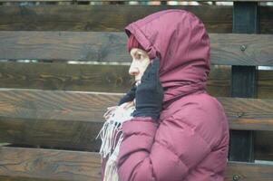 ein kaukasisch Frau im ein Burgund Jacke und ein kariert Gelb Schal Abdeckungen ihr Kopf und Gesicht mit ein Kapuze von das Wetter und Wind. Wetter Schutz. foto