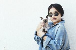 jung attraktiv Frau umarmen Muschi Katze im Hände. süß und glamourös Mädchen im modisch Sonnenbrille posieren mit ihr Siamese Katze foto