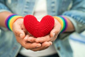 asiatische dame, die regenbogenflaggenarmbänder trägt und rotes herz hält, symbol des lgbt-stolzmonats, feiert jährlich im juni sozial für schwule, lesbische, bisexuelle, transgender, menschenrechte. foto