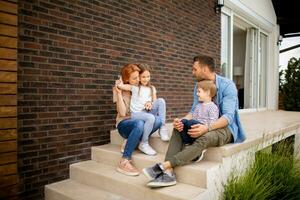 Familie mit ein Mutter, Vater, Sohn und Tochter Sitzung draußen auf das Schritte von ein Vorderseite Veranda von ein Backstein Haus foto