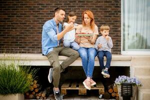 Familie mit ein Mutter, Vater, Sohn und Tochter Sitzung draußen auf Schritte von ein Vorderseite Veranda von ein Backstein Haus und Essen Erdbeeren foto