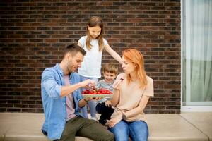 Familie mit ein Mutter, Vater, Sohn und Tochter Sitzung draußen auf Schritte von ein Vorderseite Veranda von ein Backstein Haus und Essen Erdbeeren foto