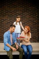 Familie mit ein Mutter, Vater und Tochter Sitzung draußen auf Schritte von ein Vorderseite Veranda von ein Backstein Haus und Essen Erdbeeren foto