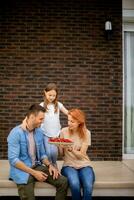 Familie mit ein Mutter, Vater und Tochter Sitzung draußen auf Schritte von ein Vorderseite Veranda von ein Backstein Haus und Essen Erdbeeren foto