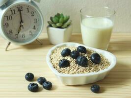 gesund Frühstück mit Hafer oder Granola im Herz gestalten Schüssel, frisch Blaubeeren, Milch und Weiß Alarm Uhr 7 bin auf hölzern Tisch. gesund Frühstück Konzept. foto