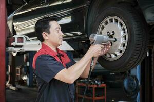 einer Experte asiatisch männlich Automobil Mechaniker Techniker ist Schrauben Auto Rad Nüsse auf Heben mit bohren Schlüssel zum Reparatur beim Garage. Fahrzeug Instandhaltung Bedienung funktioniert Industrie Besetzung Geschäft Arbeitsplätze. foto