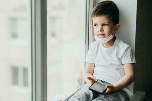 ein klein Kind im ein medizinisch Maske sitzt unter Quarantäne gestellt beim Zuhause auf ein Fenster mit ein Telefon im seine Hände.Prävention von Coronavirus und covid - - 19 foto