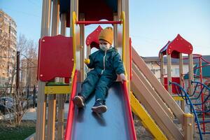 ein Kind im ein medizinisch Maske Theaterstücke auf das Spielplatz während das Pandemie von Coronavirus und covid - - 19 foto