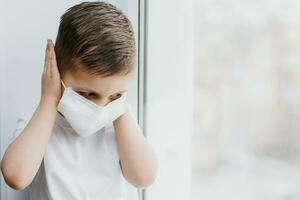 ein Kind im ein medizinisch Maske ist Sitzung beim Zuhause im Quarantäne weil von Coronavirus und covid -19 und sieht aus aus das Fenster. foto