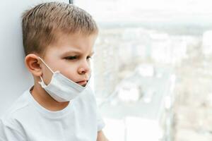 ein Kind im ein medizinisch Maske ist Sitzung beim Zuhause im Quarantäne weil von Coronavirus und covid -19 und sieht aus aus das Fenster. foto