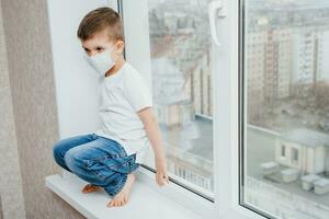 ein Kind im ein medizinisch Maske ist Sitzung beim Zuhause im Quarantäne weil von Coronavirus und covid -19 und sieht aus aus das Fenster. foto