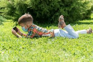 ein Kind im ein medizinisch Maske Lügen auf das Gras und sieht aus im das Telefon Cartoons im das Sommer- beim Sonnenuntergang. Kind mit ein Handy, Mobiltelefon Telefon im seine Hände. Verhütung gegen Coronavirus covid-19 während ein Pandemie foto