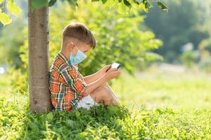 ein Kind im ein medizinisch Maske sitzt auf das Gras und sieht aus im das Telefon Cartoons im das Sommer- beim Sonnenuntergang. Kind mit ein Handy, Mobiltelefon Telefon im seine Hände. Verhütung gegen Coronavirus covid-19 während ein Pandemie foto