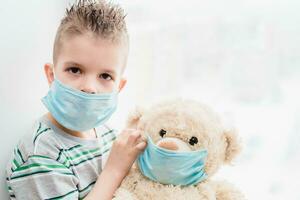 ein klein Kind mit ein Teddy Bär sitzt beim Zuhause im Quarantäne im medizinisch Masken. Verhütung von Coronavirus und covid - - 19. foto