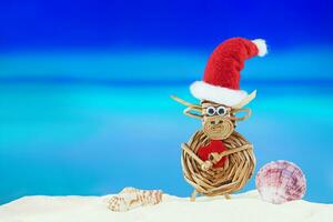Spielzeug gewebte Kuh mit Weihnachtsmann Hut mit Herz und Muscheln auf sandig Strand mit Meer. Weihnachten, Neu Jahr, Valentinstag Tag. heiß Länder. Kopieren Raum foto