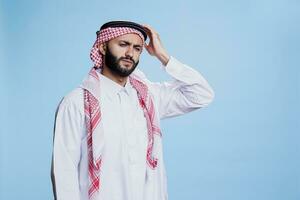 erschöpft Muslim Mann gekleidet im traditionell Kleider halten Kopf im Schmerzen während Leiden von Migräne. müde arabisch Person tragen islamisch thobe und Kopftuch haben Kopfschmerzen foto