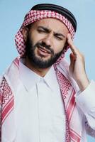 Muslim Mann zeigen Zeichen von Migräne und suchen beim Kamera mit Beschwerden Ausdruck. arabisch Person tragen traditionell Kleider haben Kopfschmerzen und Reiben Tempel Studio Porträt foto