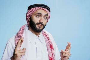 arabisch Mann tragen traditionell thobe und Kopftuch Kreuzung Finger zum gut Glück. Person gekleidet im islamisch kulturell Kleider zeigen abergläubisch Geste, Bitten zum Erfolg foto