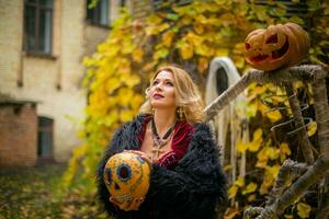 schön Frau im ein Hexe Kostüm hält ein Kürbis gemalt im Ack-o-Laternen Stil. dämpfen Kürbis im das Dekor zum Halloween. Zauberin auf ein Hintergrund von Geister. foto