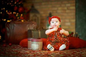 wenig Mädchen im rot Kleid gegen Hintergrund von Weihnachten Baum hält Weihnachten Girlande im ihr Hände. Baby 6 Monat alt feiert Weihnachten. foto