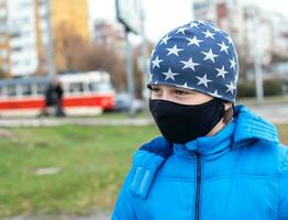 Junge im schwarz Gesicht Maske auf Straße in der Nähe von Straßenbahn im Ukraine. Coronavirus Epidemie. Gesicht Maske gegen Virus, krank, Epidemie, Grippe. Coronavirus. Gesicht Maske zum Schutz Coronavirus Ausbruch foto