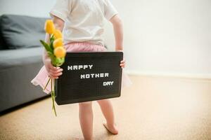 wenig Mädchen hält ein Strauß von Gelb Tulpen und ein Zeichen Das sagt glücklich Mutter Tag. Kind gibt Blumen zu Mutter zum Urlaub. foto