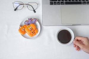 Hand hält Kaffeetasse und isst Halloween-Kekse während der Verwendung von Computer-Laptop. Frohes Halloween, Online-Shopping, Hallo Oktober, Herbst Herbst, Fest, Party und Urlaubskonzept foto