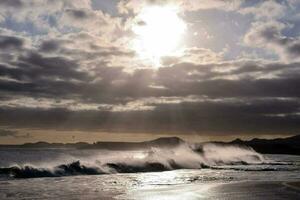 das Sonne scheint durch das Wolken Über Wellen abstürzen auf ein Strand foto