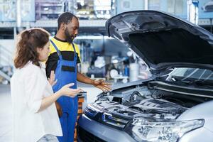 afrikanisch amerikanisch Mechaniker Portion Klient mit Auto Instandhaltung im Auto Reparatur Geschäft. Mitarbeiter im Garage Einrichtung suchen Über Automobil Teile mit Frau, Ausbessern ihr Fahrzeug Motor während Inspektion foto