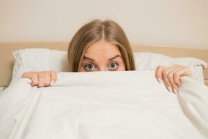 Frau mit schön Blau Augen Verlegung im Bett und spähen hinter Bettdecke foto