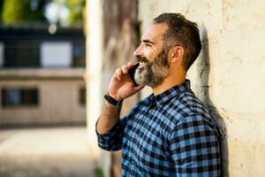 modern Geschäftsmann mit Bart mit Handy, Mobiltelefon Telefon während Stehen im Vorderseite von Mauer draussen foto