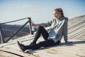 sportlich Frau ruhen nach Übung und Trinken Wasser während Sitzung auf das alt Brücke im Natur foto