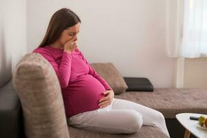 schwanger Frau fühlt sich krank während während Sitzung auf Sofa beim ihr heim. foto