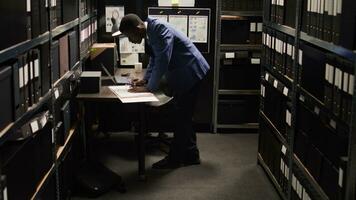 afrikanisch amerikanisch Polizist vorsichtig Analysen Material, tut Aufzeichnungen sucht, und sammelt Daten von Regale und Laptop. männlich Polizei Offizier Stehen, suchen durch Fall Dateien auf Schreibtisch im Büro. foto