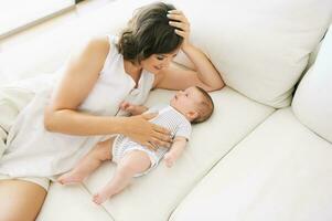 Innen- Porträt von glücklich jung Mutter spielen mit bezaubernd wenig Baby, Lügen auf Weiß Couch foto