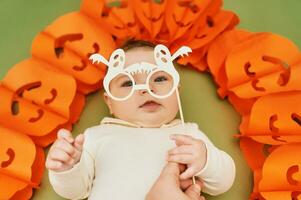 Halloween Porträt von bezaubernd Baby Lügen auf Grün Hintergrund Nächster zu Kürbis Girlande, halten Geist Papier Brille zum Foto Stand