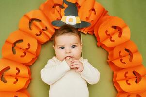 Halloween Porträt von bezaubernd Baby Lügen auf Grün Hintergrund Nächster zu Kürbis Girlande, Hexe Papier Hut festlich Party Requisiten foto