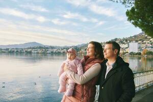 draussen Porträt von glücklich jung Paar mit bezaubernd Baby Mädchen genießen nett Aussicht von Winter See Genf oder lac leman, Montreux, Schweiz foto