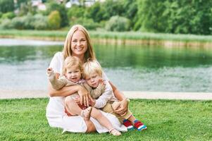 draussen Porträt von glücklich jung Mutter mit zwei schön Kinder spielen durch See oder Fluss auf ein nett Sommer- Tag foto